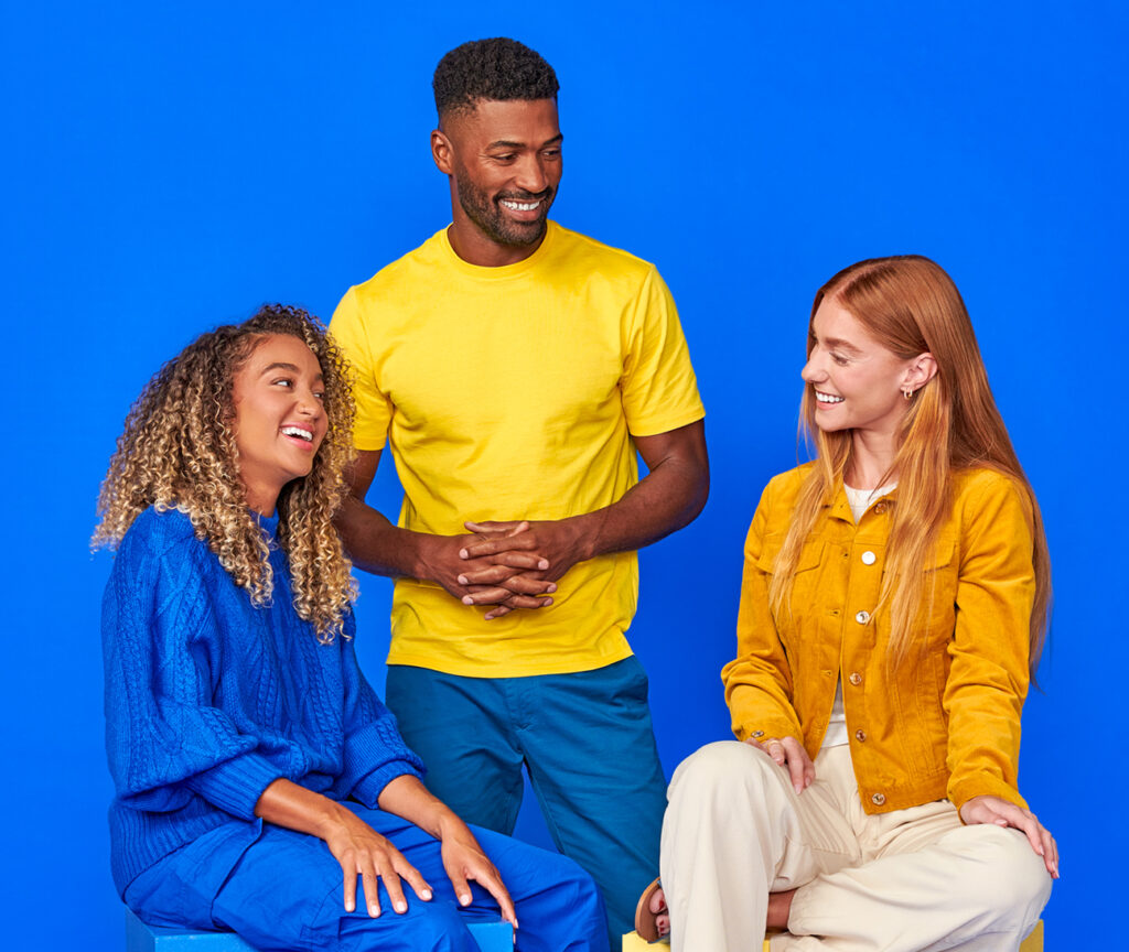 Two women sitting and a man standing in the middle against a blue background, smiling and looking at the each other.