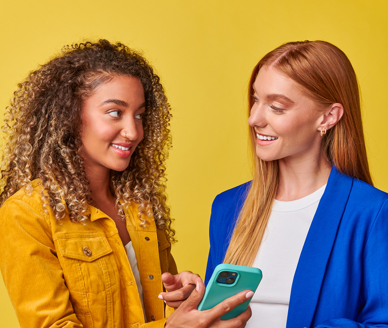Two women stand against a yellow background, smiling and looking at each other. One holds a smartphone.